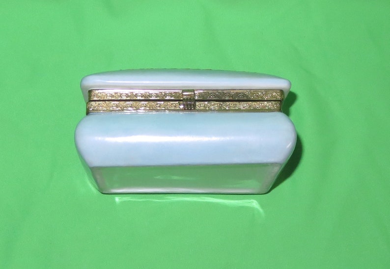 May include: A white porcelain trinket box with a gold-colored hinged lid. The box has a rectangular shape with rounded edges.