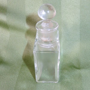 May include: A clear glass bottle with a round glass stopper. The bottle has a square shape and is empty.
