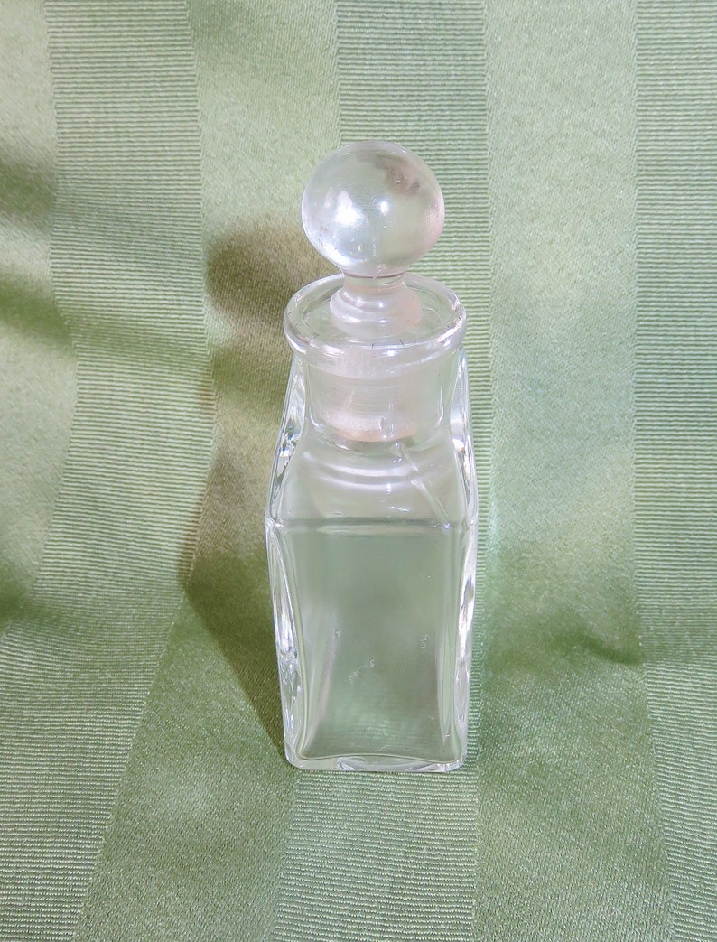 May include: A clear glass bottle with a round glass stopper. The bottle has a square shape and is empty.