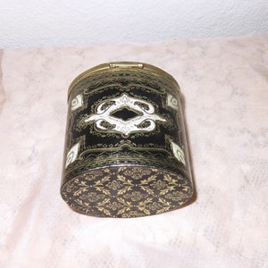 May include: A black cylindrical container with a gold lid and intricate gold and white designs. The container has a floral pattern on the bottom.