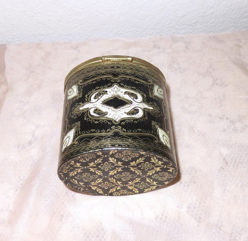 May include: A black cylindrical container with a gold lid and intricate gold and white designs. The container has a floral pattern on the bottom.