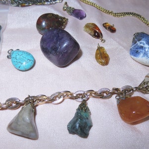 May include: A gold chain necklace with various gemstone pendants, including turquoise, amethyst, carnelian, and clear quartz. The necklace is displayed on a pink fabric background.