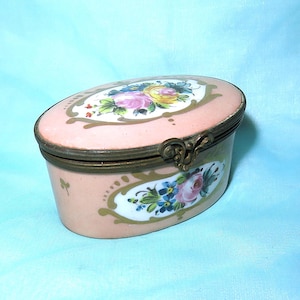 Pode incluir: Uma caixa de porcelana oval com tampa articulada. A caixa é decorada com desenhos florais pintados à mão em tons de rosa, azul e verde. A tampa tem uma moldura dourada e um desenho floral com uma rosa rosa no centro.