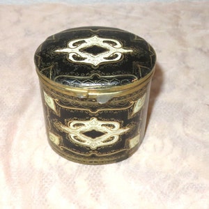 May include: A black and gold cylindrical tin with a hinged lid. The lid and sides are decorated with a white and gold geometric pattern.