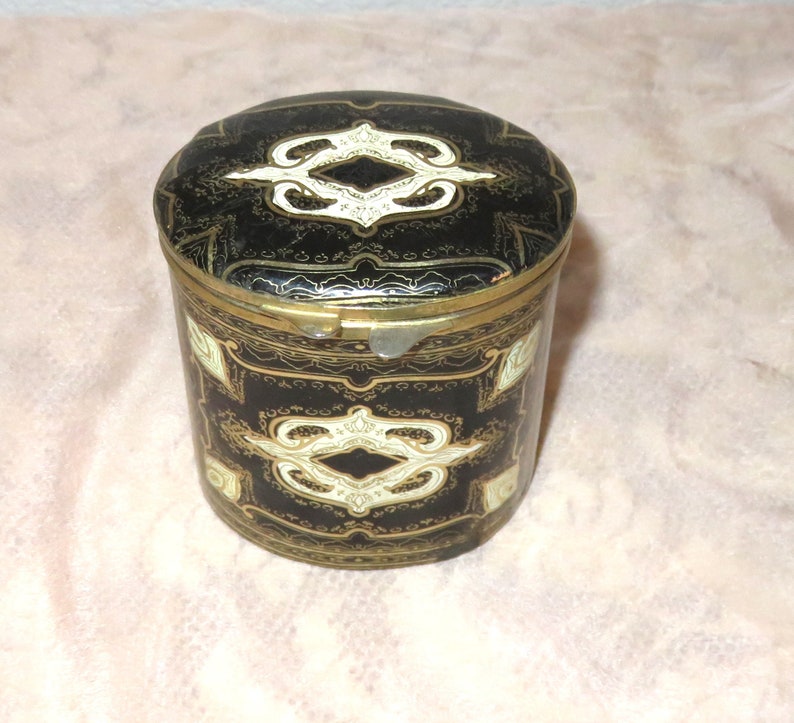 May include: A black and gold cylindrical tin with a hinged lid. The lid and sides are decorated with a white and gold geometric pattern.
