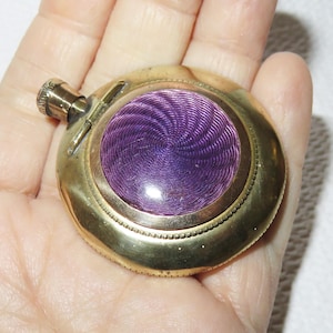 May include: A round, brass lighter with a purple enamel design. The lighter has a small, round lid and a spout on the side.