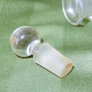 May include: A clear glass bottle stopper with a round top and a frosted, cylindrical base.