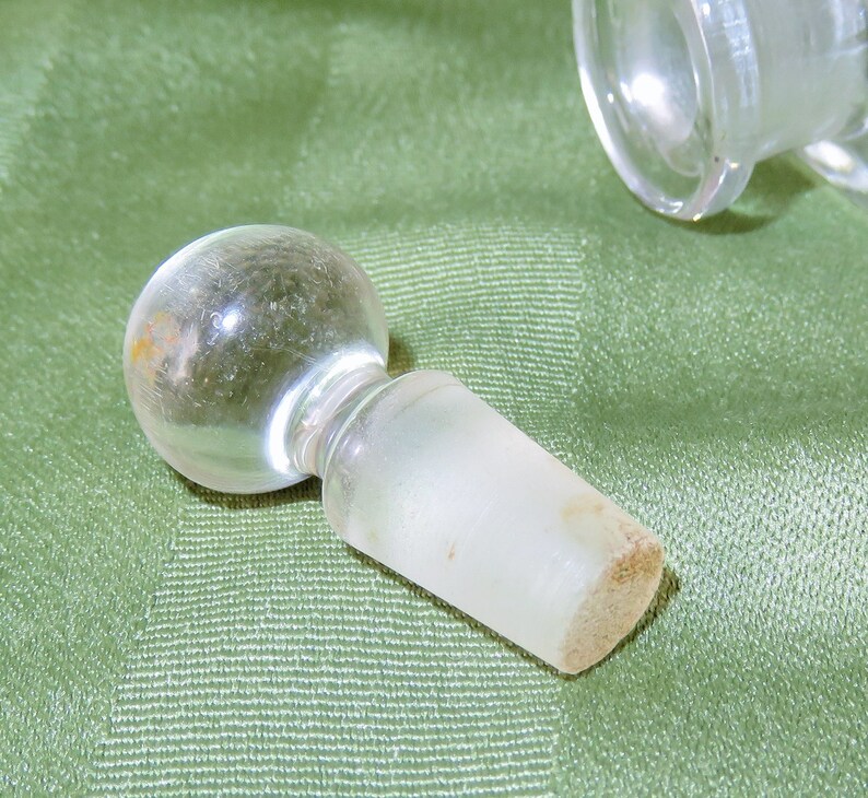 May include: A clear glass bottle stopper with a round top and a frosted, cylindrical base.
