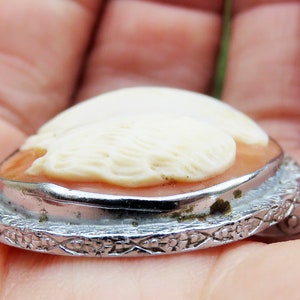 May include: A cameo pendant featuring a carved white design on a pink background, set in a silver tone metal frame with a detailed floral pattern.