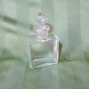 May include: A clear glass bottle with a round glass stopper. The bottle is square shaped and has a smooth surface.