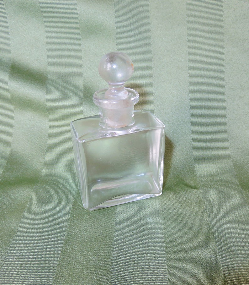 May include: A clear glass bottle with a round glass stopper. The bottle is square shaped and has a smooth surface.