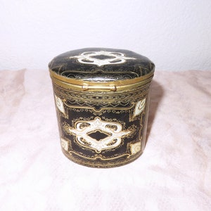May include: A black and gold cylindrical box with a hinged lid. The box is decorated with intricate white and gold patterns.