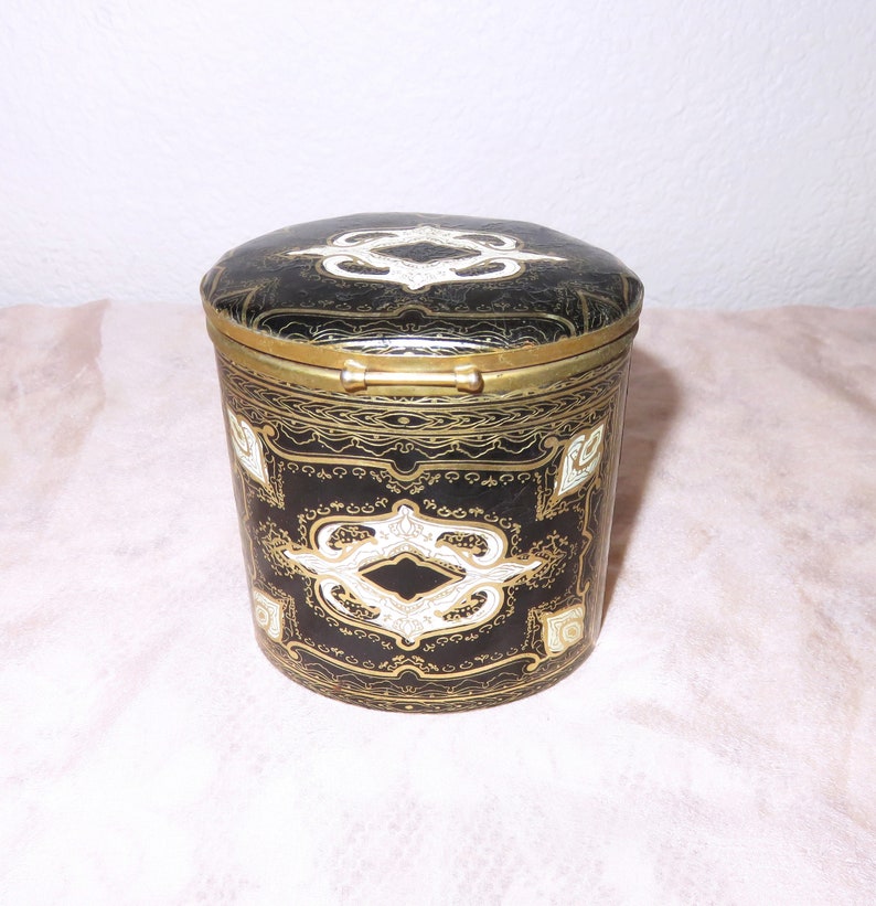 May include: A black and gold cylindrical box with a hinged lid. The box is decorated with intricate white and gold patterns.