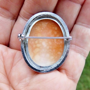 May include: A silver cameo brooch with a delicate, oval-shaped shell cameo. The back of the brooch features a silver pin and a hallmark.