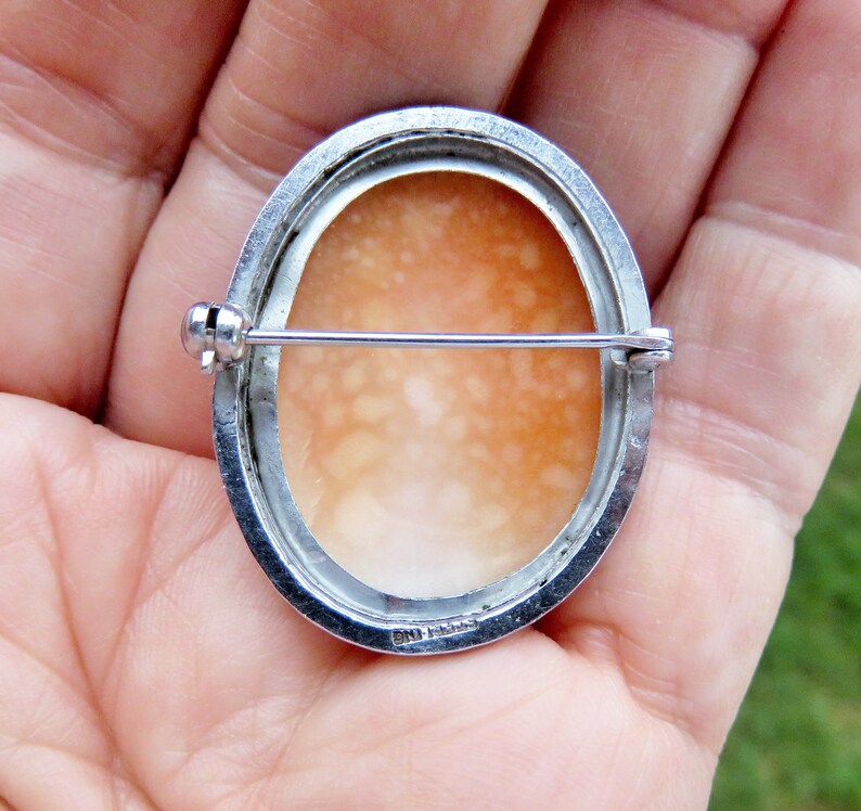 May include: A silver cameo brooch with a delicate, oval-shaped shell cameo. The back of the brooch features a silver pin and a hallmark.