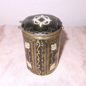 May include: A black cylindrical container with a gold and white patterned design. The container has a hinged lid with a decorative design.