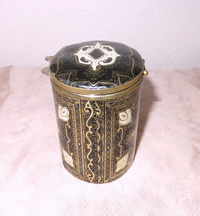 May include: A black cylindrical container with a gold and white patterned design. The container has a hinged lid with a decorative design.