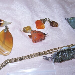 May include: A collection of seven gemstone pendants, some wrapped in wire, with various colors including brown, orange, green, and white. The pendants are displayed on a light pink fabric surface.
