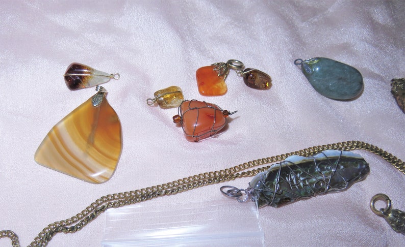 May include: A collection of seven gemstone pendants, some wrapped in wire, with various colors including brown, orange, green, and white. The pendants are displayed on a light pink fabric surface.