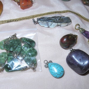 May include: A collection of gemstone pendants, including green jade, purple amethyst, turquoise, and amber. The pendants are displayed on a white surface with a gold chain.