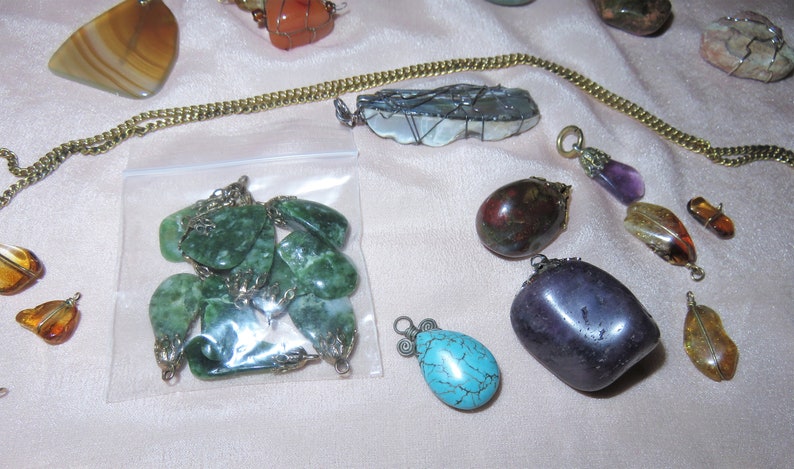 May include: A collection of gemstone pendants, including green jade, purple amethyst, turquoise, and amber. The pendants are displayed on a white surface with a gold chain.