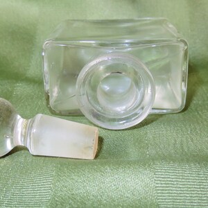 May include: A clear glass bottle with a glass stopper. The bottle has a square shape and the stopper is round. The bottle is empty.