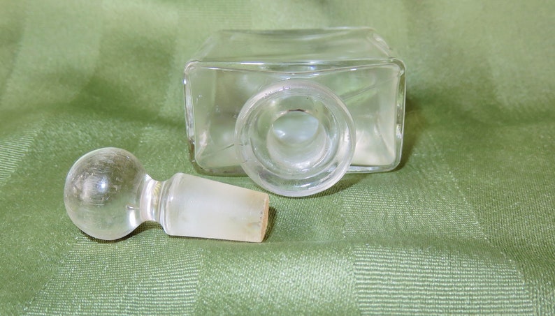 May include: A clear glass bottle with a glass stopper. The bottle has a square shape and the stopper is round. The bottle is empty.