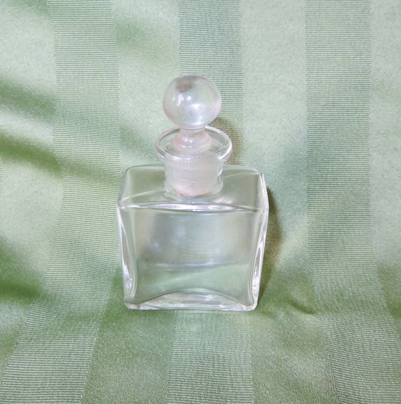 May include: A clear glass bottle with a round glass stopper. The bottle is square shaped and has a smooth surface.