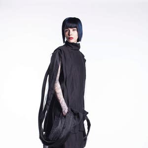 K&ouml;nnte beinhalten: Eine Person tr&auml;gt ein schwarzes Outfit mit einem langen, flie&szlig;enden Rock und einem hochgeschlossenen Oberteil. Die Person hat kurzes, dunkelblaues Haar und tr&auml;gt schwarze Turnschuhe.