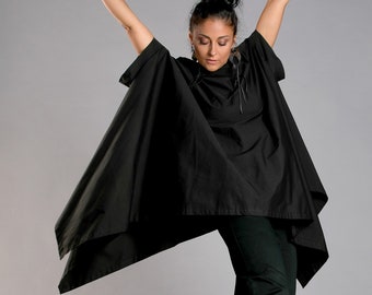 May include: A woman wearing a black, loose-fitting top with a long, flowing hem and black pants. She is wearing colorful sneakers and has her arms raised above her head.