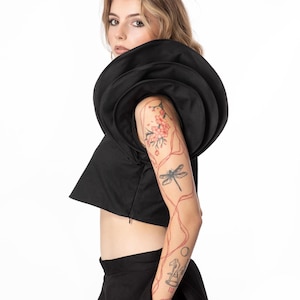 May include: A black cropped top featuring a prominent, multi-layered circular ruffle detail around the shoulder and neckline, paired with a matching black skirt. The top has a side zipper closure and a structured, sleeveless design.