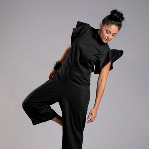 May include: A woman wearing a black jumpsuit with ruffled sleeves and green heels. She is standing on one leg with her other leg bent at the knee.