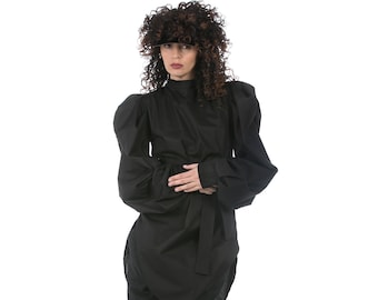 Op de afbeelding: Een zwarte outfit met een top met hoge halslijn en volumineuze mouwen, een wikkelrok en een cropped broek. De outfit is in de taille vastgemaakt met een bijpassende riem. Witte sneakers maken de look compleet.