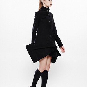 May include: A woman wearing a black coat with a high collar and a button closure. She is wearing black knee-high socks and black platform sandals with a wooden heel.