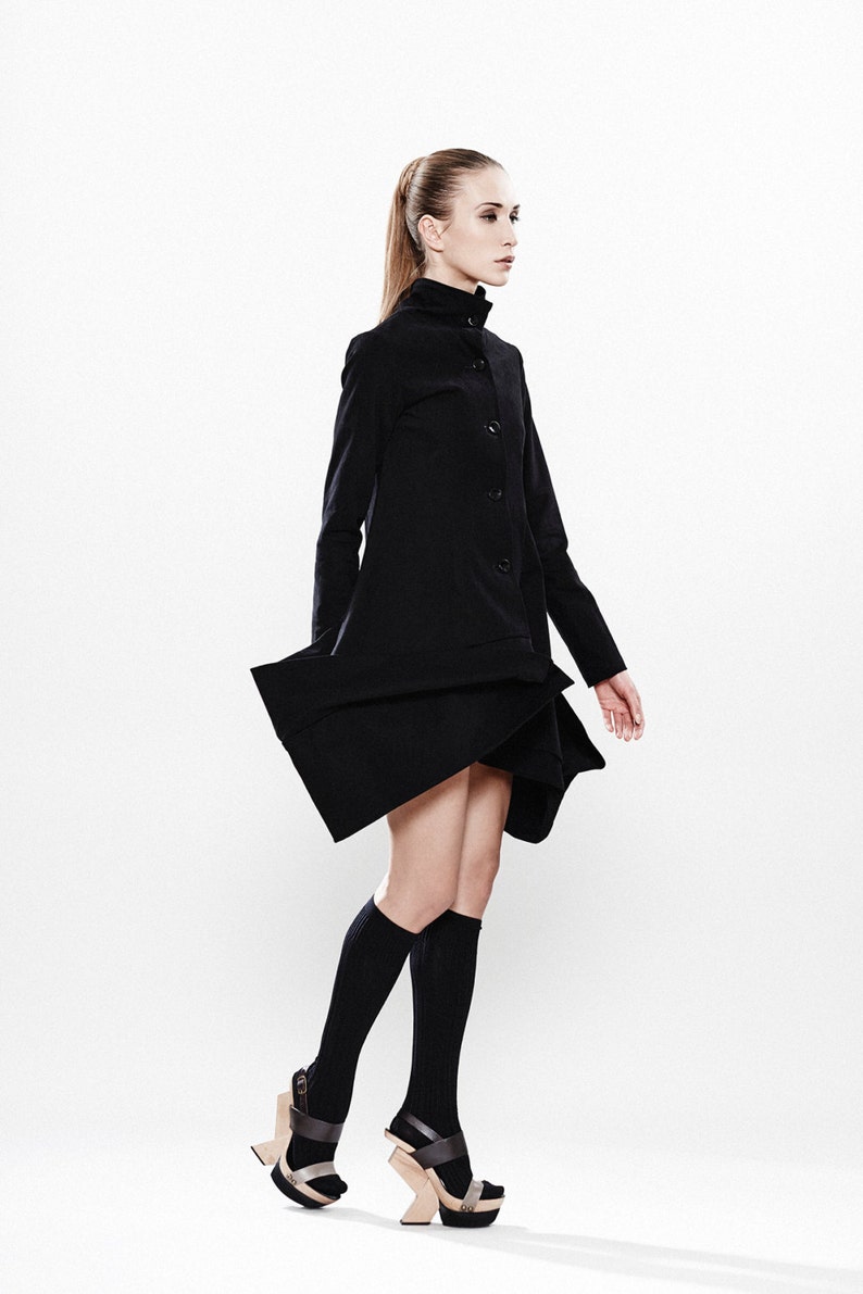 May include: A woman wearing a black coat with a high collar and a button closure. She is wearing black knee-high socks and black platform sandals with a wooden heel.