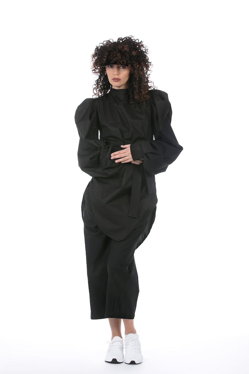 May include: A black outfit featuring a high-neck top with voluminous sleeves, a wrap-style skirt, and cropped pants. The outfit is cinched at the waist with a matching belt. White sneakers complete the look.
