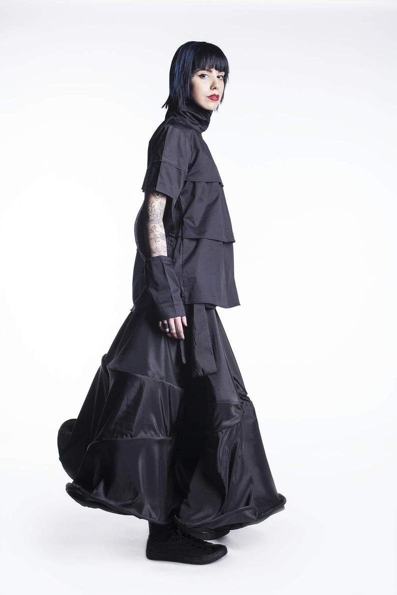 May include: A woman wearing a black layered top and a black skirt with multiple tiers. She is wearing black sneakers and has dark blue hair.