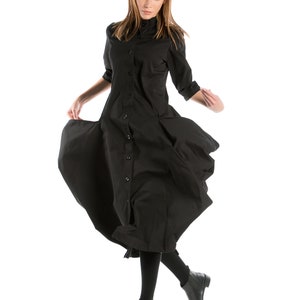 K&ouml;nnte beinhalten: Eine Frau in einem schwarzen Knopfkost&uuml;m mit langen &Auml;rmeln und schwarzen Stiefeletten. Das Kleid hat einen flie&szlig;enden Rock und die Frau ist in der Luft, mit gespreizten Beinen.