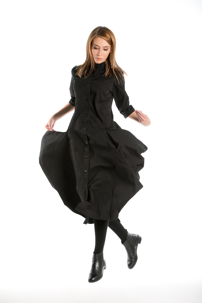 K&ouml;nnte beinhalten: Eine Frau in einem schwarzen Knopfkost&uuml;m mit langen &Auml;rmeln und schwarzen Stiefeletten. Das Kleid hat einen flie&szlig;enden Rock und die Frau ist in der Luft, mit gespreizten Beinen.