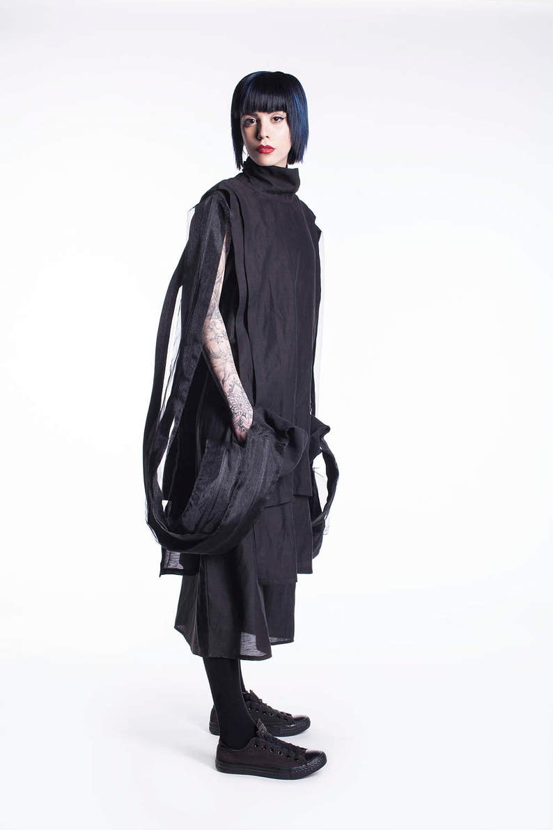 K&ouml;nnte beinhalten: Eine Person tr&auml;gt ein schwarzes Outfit mit einem langen, flie&szlig;enden Rock und einem hochgeschlossenen Oberteil. Die Person hat kurzes, dunkelblaues Haar und tr&auml;gt schwarze Turnschuhe.
