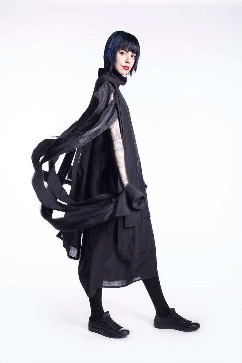 K&ouml;nnte beinhalten: Eine Frau tr&auml;gt ein schwarzes, flie&szlig;endes Kleid mit hohem Kragen und langen &Auml;rmeln. Das Kleid hat ein einzigartiges Design mit Stoffschichten, die einen dramatischen Effekt erzeugen. Sie tr&auml;gt schwarze Strumpfhosen und schwarze Turnschuhe.