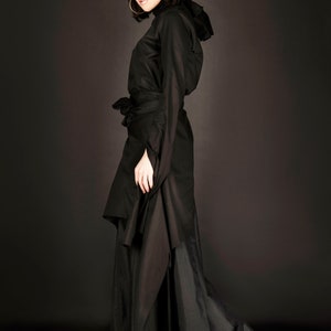 May include: A woman wearing a black long-sleeved dress with a high neck and a belt. The dress has a flowing skirt and is made of a soft, drapey fabric.