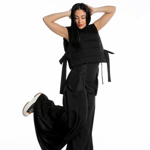 May include: A black sleeveless vest with tie closures, paired with wide-leg black pants. The outfit is completed with a pair of white and brown sneakers. The model is posing with their arms above their head.