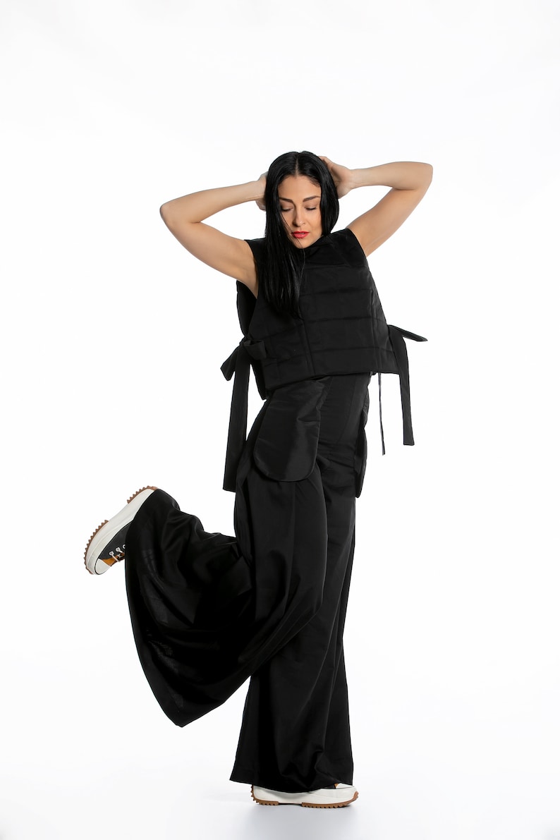May include: A black sleeveless vest with tie closures, paired with wide-leg black pants. The outfit is completed with a pair of white and brown sneakers. The model is posing with their arms above their head.