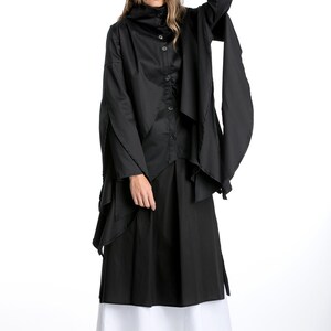 May include: A black long-sleeved shirt with a high collar and a layered design. The shirt is worn with a black skirt and white underskirt. The model is wearing black shoes.