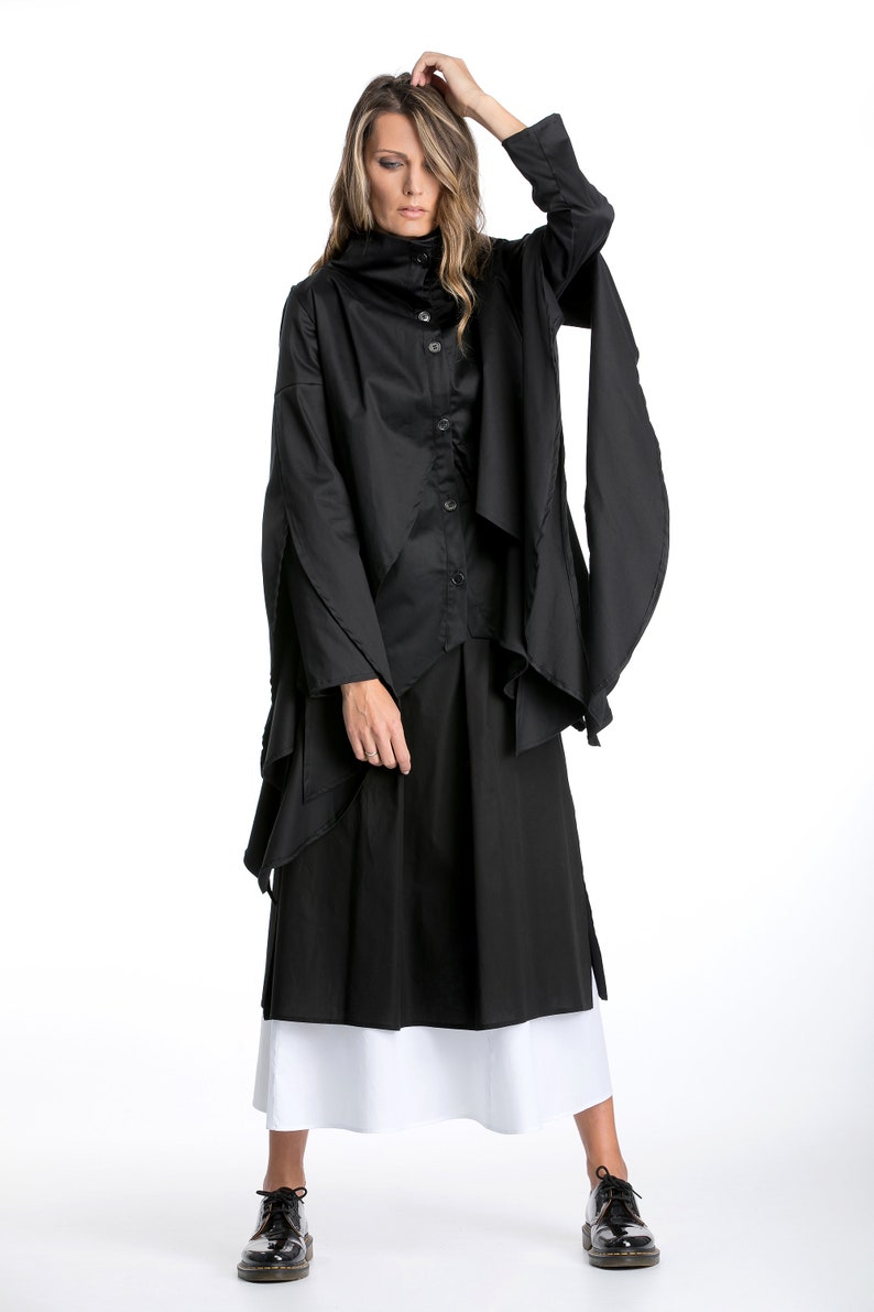 May include: A black long-sleeved shirt with a high collar and a layered design. The shirt is worn with a black skirt and white underskirt. The model is wearing black shoes.