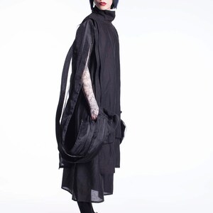 K&ouml;nnte beinhalten: Eine Frau tr&auml;gt ein schwarzes, mehrschichtiges Kleid mit einem langen, flie&szlig;enden Schal. Sie hat kurzes, dunkelblaues Haar und tr&auml;gt schwarze Turnschuhe.