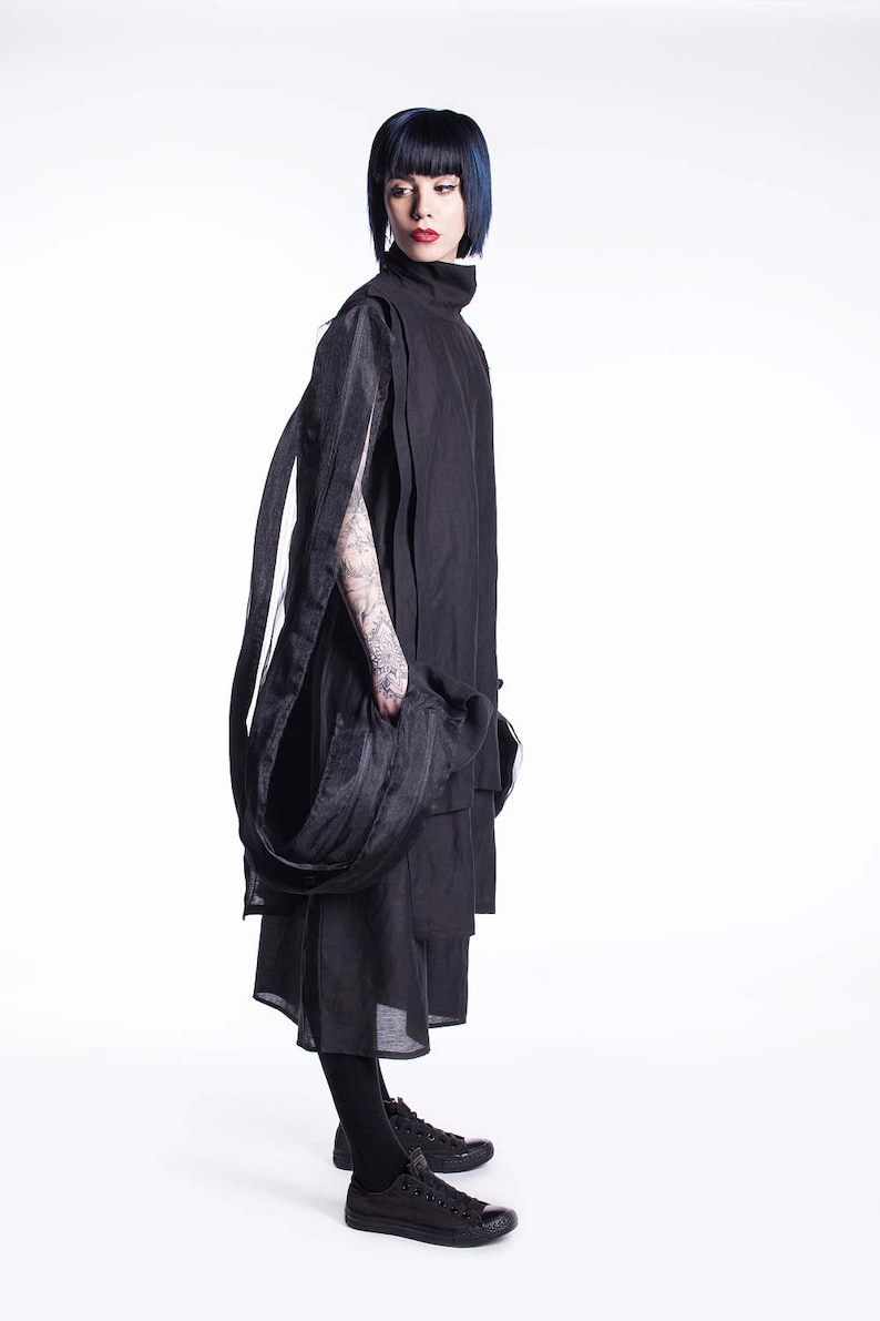 K&ouml;nnte beinhalten: Eine Frau tr&auml;gt ein schwarzes, mehrschichtiges Kleid mit einem langen, flie&szlig;enden Schal. Sie hat kurzes, dunkelblaues Haar und tr&auml;gt schwarze Turnschuhe.