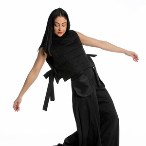 May include: Black sleeveless vest with a quilted design, paired with wide-leg pants. The vest features side ties and pockets. The model is wearing white sneakers with a brown sole. The outfit is all black.