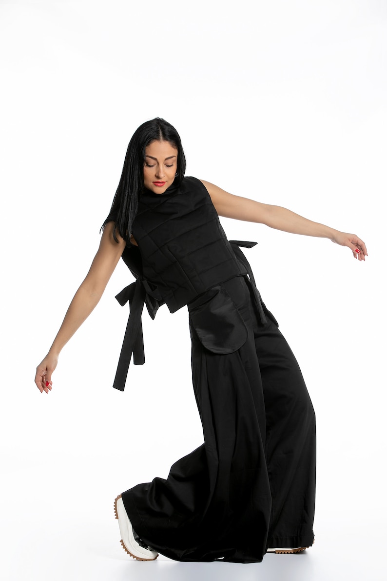 May include: Black sleeveless vest with a quilted design, paired with wide-leg pants. The vest features side ties and pockets. The model is wearing white sneakers with a brown sole. The outfit is all black.